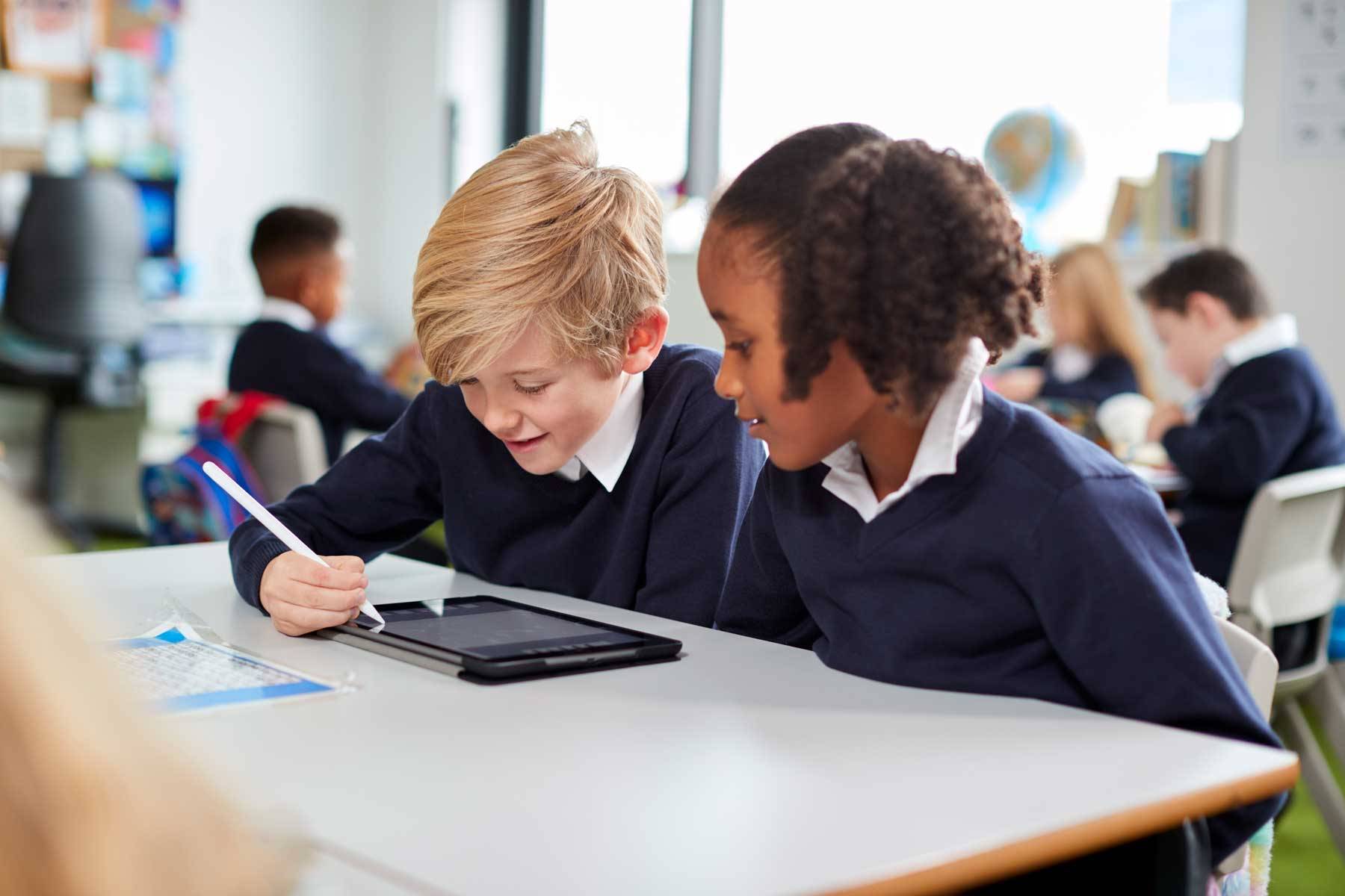 two-primary-school-kids-sitting-together-desk-classroom-using-tablet-computer-stylus-close-up image